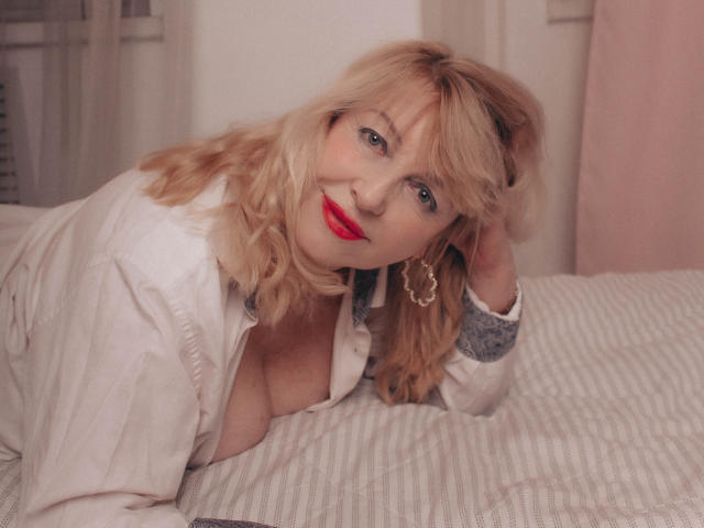 SamanthaLala with blonde hair, red lipstick, and hoop earrings, lying on a bed, wearing a white shirt, exposing her large, bare tits, smiling seductively at the camera.