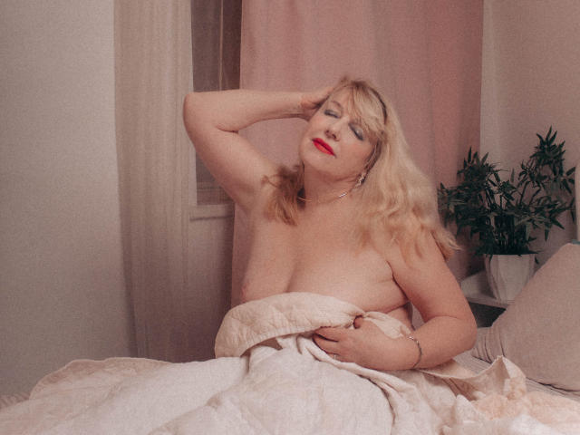 SamanthaLala mit blonden Haaren, rotem Lippenstift und heller Haut, sitzt oben ohne und bedeckt ihre Brüste mit einer weißen Decke, eine Hand in ihrem Haar, im Hintergrund eine pralle, grüne Pflanze..