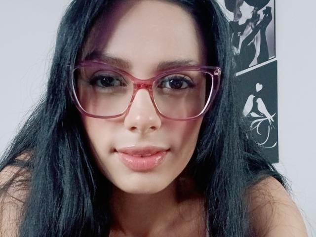 AnaisHilton with long black hair, pink glasses, and shiny pink lips, looking straight at the camera, her nose slightly upturned, wearing a sleeveless top, her tits barely visible.