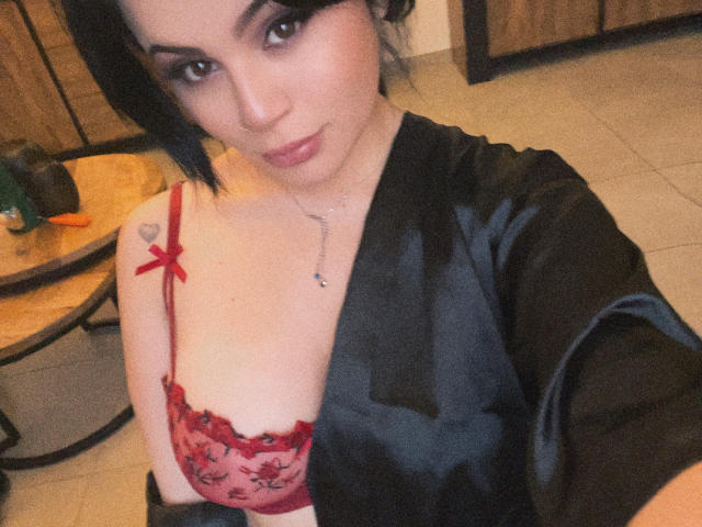 ShantallParis with black hair, wearing a black satin robe and red lace bra, showing small tits with a red ribbon and heart tattoo on her right shoulder.