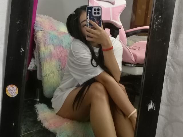 OdettePrincess with long black hair, sitting on a fluffy, multi-colored chair, wearing a white t-shirt, taking a mirror selfie, showing her bare ass and legs, holding a phone.