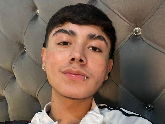 ImJuanda has short black hair, brown eyes, and a small nose ring. He's wearing a white shirt with black stripes, and his chin is slightly shiny.