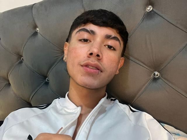 ImJuanda, with short black hair and light skin, wearing a white jacket, has a small nose ring and is lying on a tufted gray headboard, looking at the camera.