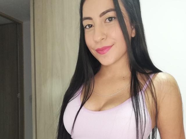 MilauraSwt with long black hair, pink lipstick, and a cute smile, wearing a pink tank top that shows her big tits. She's got a slim build and a necklace.