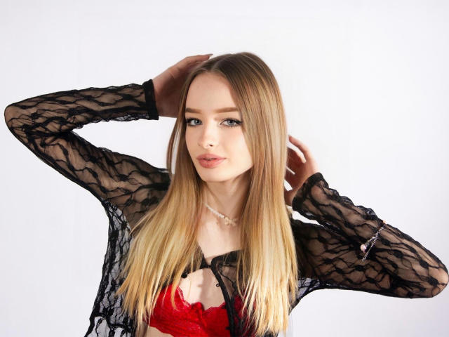 LilaFantasy, con cabello rubio largo, vestida con una blusa negra de encaje y un sujetador rojo de encaje, con los brazos levantados, mirando a la cámara, fondo blanco, expresión suave, maquillaje sutil, complexión delgada..