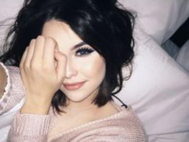 CarlaLow, com cabelos escuros e ondulados, um suéter rosa claro e uma mão cobrindo o olho, sorrindo sutilmente. Ela tem cílios longos, lábios rosados e um visual adorável e sexy..