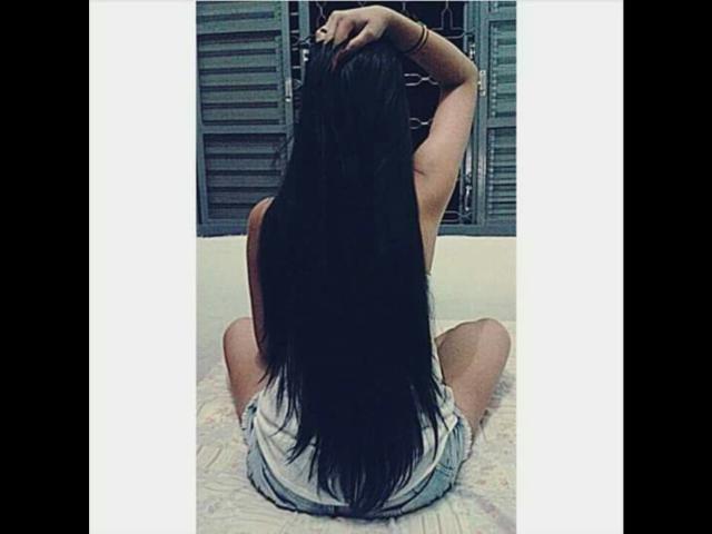 CarlaLow, com cabelos longos e pretos, de costas para a câmera, sentada na cama, com a mão no cabelo, vestindo uma blusa branca e shorts azuis, pernas dobradas, bumbum visível, pulseira no braço..