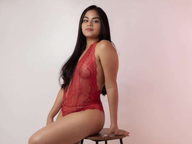 DanaCami with long black hair, wearing a red lace bodysuit, sitting on a stool, showing her tits and pussy through sheer fabric, with a confident smile.