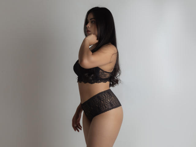 DanaCami with long black hair, wearing black lace bra and high-waisted panties, standing side-profile, hand near mouth, showing her ass and small tits.