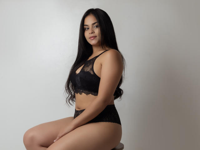 DanaCami with long black hair, wearing black lace bra and panties, sitting, showing her ass, tits in bra, and a slight smile, in a simple, light-colored room.