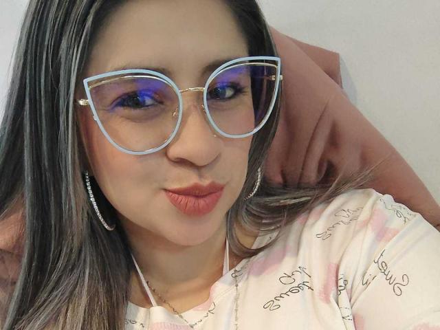 MelindaBoobs with blue-tinted glasses, pouty pink lips, and long brown hair, wearing a pink and white patterned shirt, has her hand behind her head.