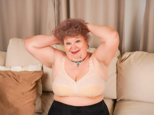 MomCum with curly pink hair, smiling, arms raised, wearing a beige bra, showing her big boobs, sitting on a beige couch, black pants, green necklace, and red lipstick.