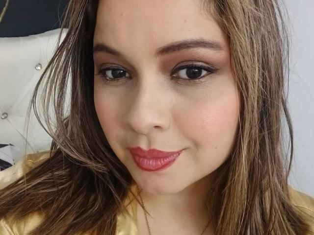 IdyllaJhones has a cute face with brown eyes, red lipstick, and brown hair, wearing a golden shirt. Her tits are not visible. She's smiling slightly.