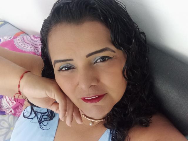 HannaSofis, con i capelli neri ricci, ombretto blu, rossetto rosso, piercing al naso, collana e braccialetto rosso, sdraiata, sorride alla fotocamera, mostrando la mano sulla guancia..