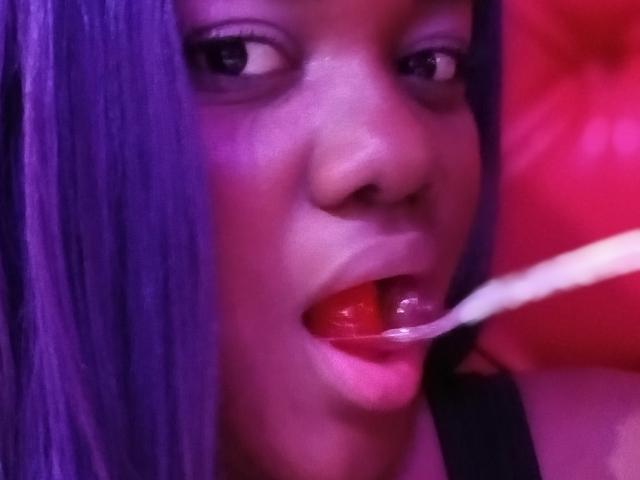 RoseBeckers with purple hair, black eyes, and glossy red lips, has a white spoon in her mouth, her tongue slightly touching the spoon's tip.