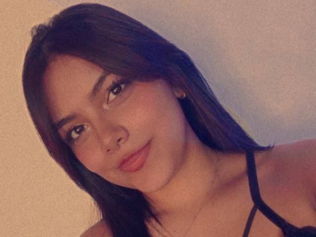 Emily Roberts tem cabelo castanho escuro, olhos castanhos e um leve sorriso com batom vermelho. Ela está vestindo uma blusa preta com alças finas, e seus pequenos seios estão levemente visíveis..