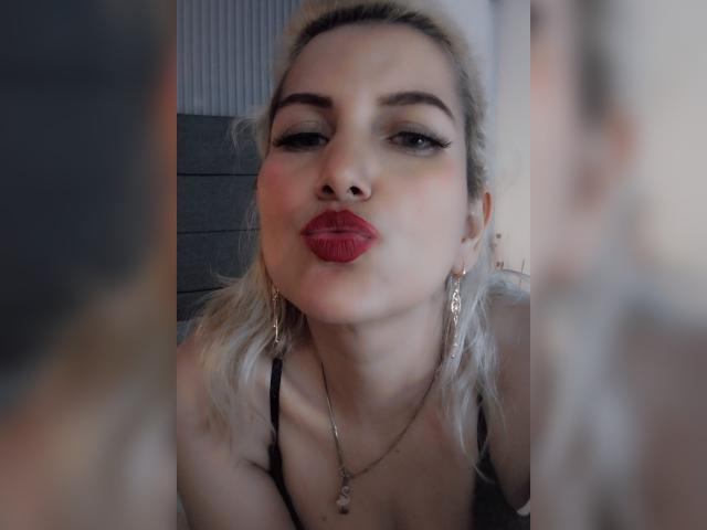 GiuliaAddams with pouty red lips, light blonde hair, and gold earrings, looking into a mirror, showing her black-strapped top and necklace, with a soft, blurry background.