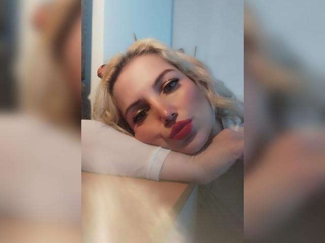 GiuliaAddams a les cheveux blonds, du rouge à lèvres et un anneau de nez, et elle est appuyée contre une surface avec une expression détendue, dans un selfie flou pris devant un miroir..