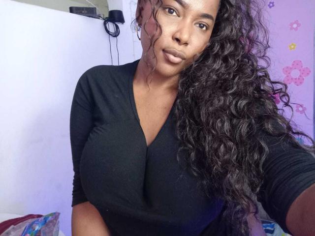 EmiraHot with big tits, dark skin, and long curly black hair wearing a black long-sleeve top, looking at the camera with a neutral expression.
