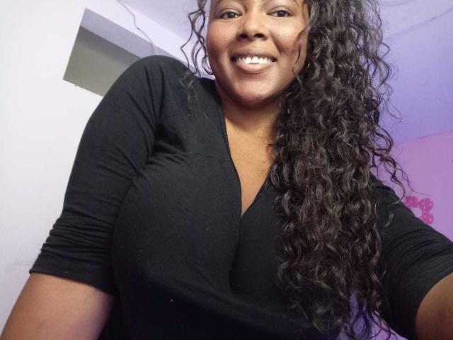 EmiraHot with dark curly hair, smiling, wearing a black V-neck shirt, showcasing large tits, standing indoors, taking a selfie, showing her teeth, and looking at the camera.