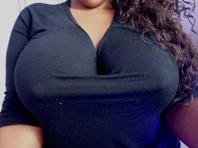 EmiraHot's large, round, black-t-shirted tits are visible, with her curly brown hair falling on the right. Her chest is prominently pushed up, showing deep cleavage.