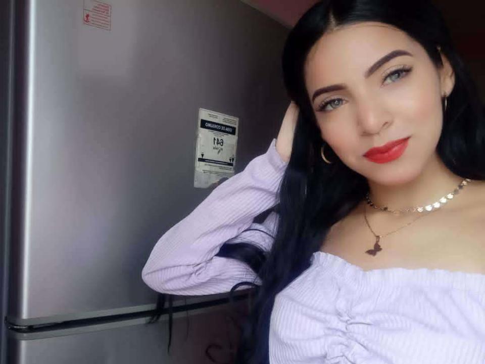 StefayGrey with long black hair, red lipstick, and a purple off-shoulder top, hand in hair, smiling, wearing a necklace with a small cross, standing by a fridge.