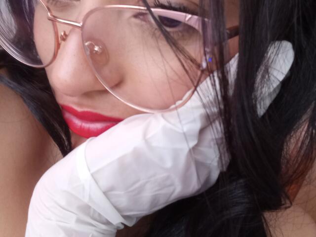 OdettePrincess with red lips, wearing pink glasses, has long black hair, white shirt, and a gloved hand near her face. Nose piercing visible. Soft, intimate close-up.