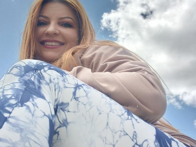 NatalyHami with blonde hair, smiling, wearing a beige hoodie and blue/white tie-dye pants, shot from low angle, showing her tits slightly, against a cloudy sky.