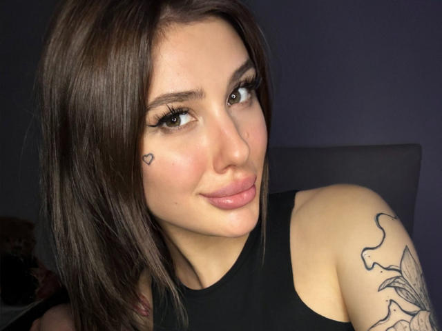 SensualMila with dark brown hair, black sleeveless top, heart tattoo on cheek, and large pink lips, showing off tattooed arm, looking to the side with a slight smile.