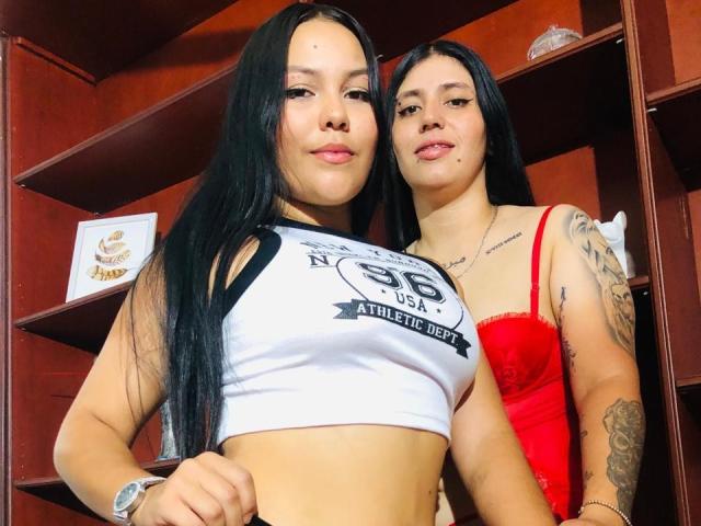 AlanaAndShaira, avec de longs cheveux noirs, portant un crop top blanc et noir qui dévoile leurs seins et un soutien-gorge en dentelle rouge, se tiennent l'une à côté de l'autre, toutes les deux souriantes, avec des tatouages visibles..