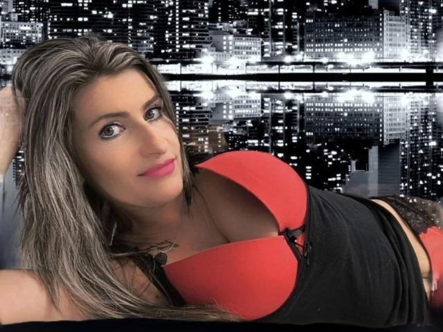 KatyStar-hot with long brown hair, pink lipstick, and red boobs in a black top, lying down, smiling, wearing black lace panties, in a city skyline photo.