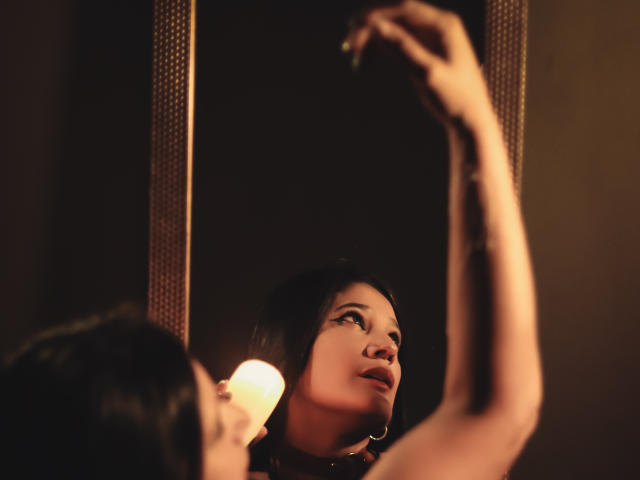PaulaCamis with dark hair, looking up at her reflection in a mirror, applying eyeliner, her arm raised, a warm light highlighting her face, her boobs slightly visible.