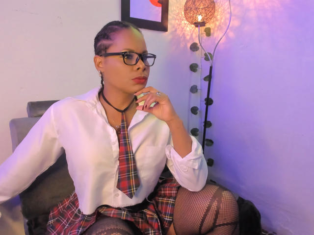 AfroditaKane mit Brille, Zöpfen, weißem Hemd, rotem Karokrawatten, Rock, Fischernetzstrümpfen und einer schwarzen Halskette, sitzend, Hand am Kinn, ernster Blick, violett-blaues Licht..