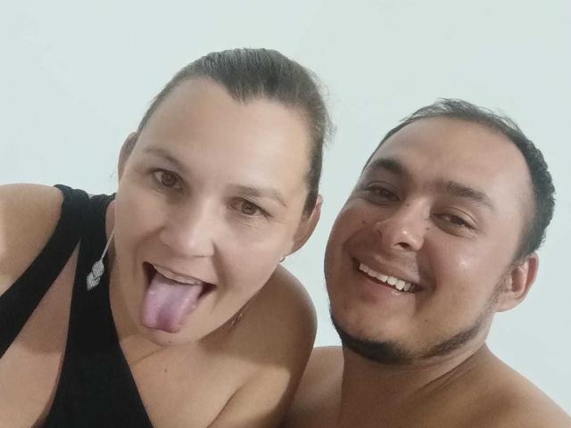 AmorDeDos69 with her tongue out, black tank top, and AmorDeDos69 shirtless, smiling, both with light skin, brown hair, and small to medium-sized tits.