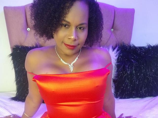 AfroditaKane con i capelli ricci, vestito di seta rossa, con spalline scoperte, collana scintillante, sorridente, sul letto, cuscini neri e pelosi, testiera rosa, luce viola, che mostra i suoi seni..