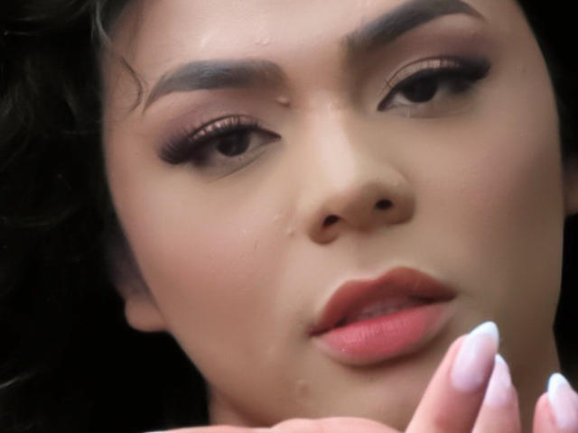 LiliithBuckland's face with glistening skin, dark eyebrows, long eyelashes, pink lips, and a slightly parted mouth. Her hand with white nails is near her chin.