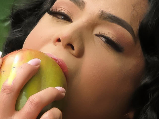 LiliithBuckland's face, eyes half-closed, biting into a green apple with her pink-lipped, manicured fingers. Her dark curly hair and brown eyebrows are visible.