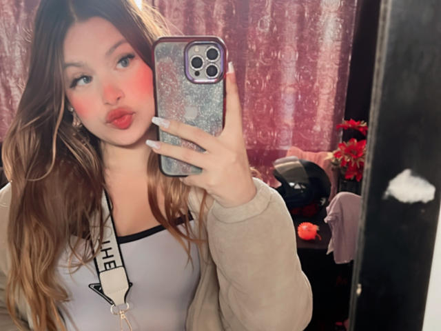 Shantal, con le guance rosse, le labbra piene e i lunghi capelli castani, sta scattando un selfie allo specchio in una stanza illuminata di rosa, indossando una felpa grigia e un canottino bianco, mostrando il seno..