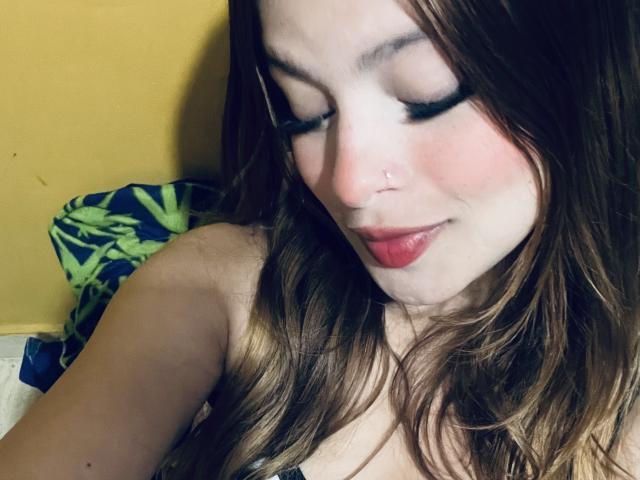 ShantalSummer with wavy brown hair, fair skin, nose piercing, red lipstick, and dark eyeliner, looking down with a slight smile, showing her bare shoulders and chest.
