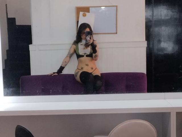 AmySpider with tattoos, wearing green bra, black arm sleeve, black thigh-highs, and black wristband, sitting on purple bench, taking mirror selfie, showing her bare ass and tits.