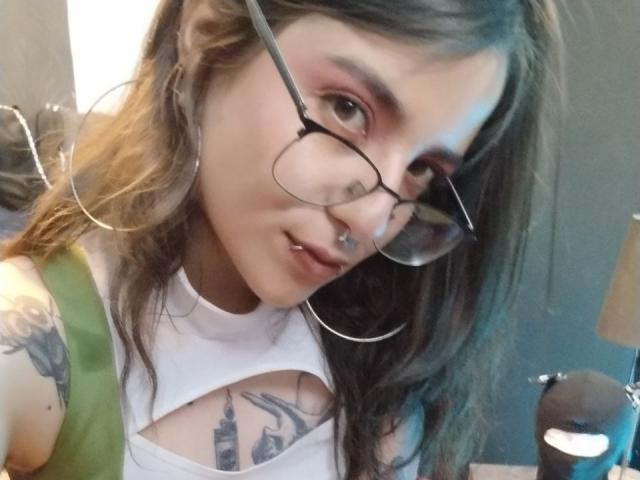 AmySpider with glasses, hoop earrings, and a white top showing her tits and a tower tattoo. She has a septum piercing and a green strap. Her makeup is pink.