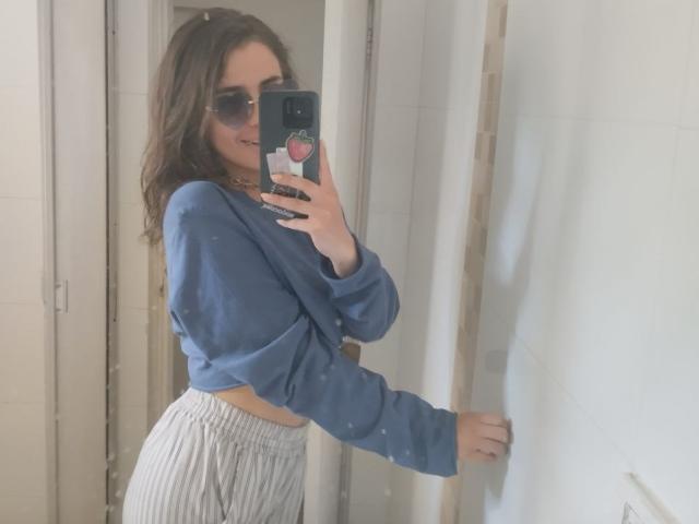 AmySpider, con cabello castaño ondulado, usando gafas de sol, un suéter azul y pantalones blancos de talle alto, está tomando una selfie con un teléfono que tiene un soporte rosa..