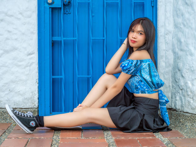MidnightHarpy with long black hair, blue off-shoulder top, black skirt, black sneakers, sitting against blue door, hand on cheek, legs extended, white textured wall behind.