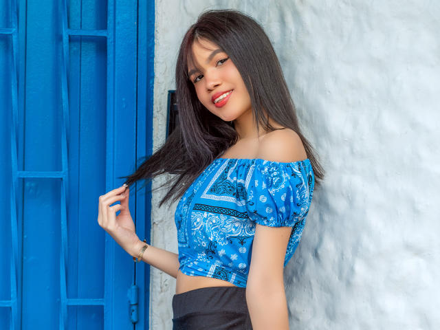 MidnightHarpy has long black hair, wearing a blue off-shoulder crop top with a pattern, black skirt, smiling with red lipstick, holding her hair, standing next to a blue door.