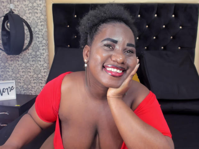 Dilay with dark skin, curly black hair, and red lipstick, smiling, head resting on hand, wearing a red off-shoulder top, exposing her large boobs.