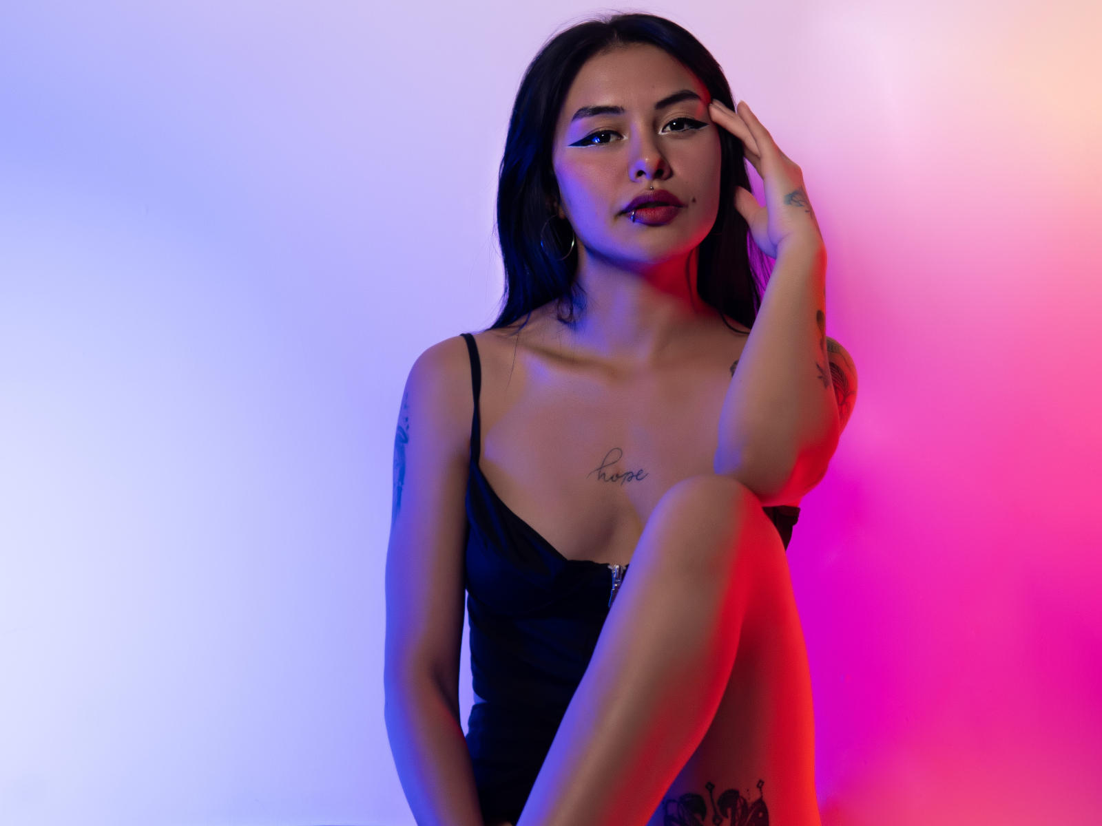 ArianaJoness with dark hair, wearing a black spaghetti strap top, sitting with knee up, hand touching face, tattoos visible, pink and blue lighting, looking at camera, sultry expression.