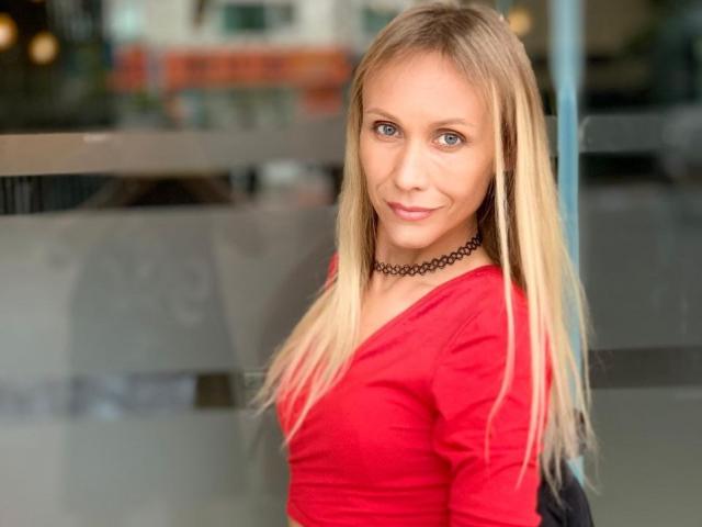 EmieFlower has blonde hair, blue eyes, and a cute smile. She's wearing a red crop top, white pants, black choker, and a black backpack. Her tits are small.