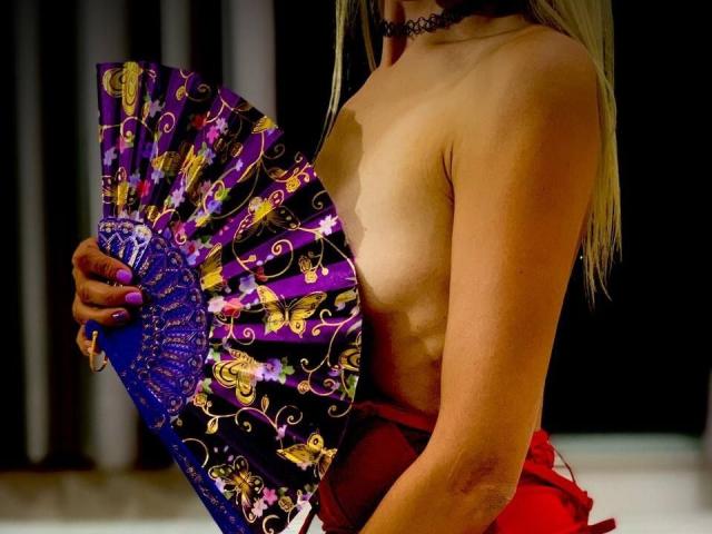 EmieFlower with a purple and gold floral fan, covering her tits, wearing a red skirt and black choker, has blonde hair, and a tan skin tone, in a dark setting.