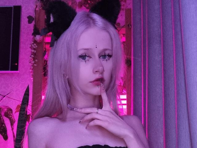 ExcitedBlizzard with pale blonde hair, black cat ears, blue eyes, and a finger near her pink-lipsticked mouth, wearing a black strap, under pink neon lighting.