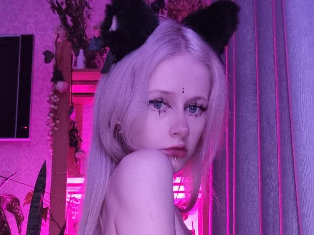 ExcitedBlizzard with pale skin, platinum blonde hair, black cat ears, blue eyes, and a nose ring, looking over her shoulder, pink neon light, and a plant in the background.